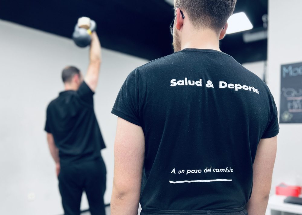 Imagen de un educador físico deportivo de espaldas con el lema de FIX "A un paso del cambio" supervisando a un usuario con kettlebell en nuestro servicio personalizado